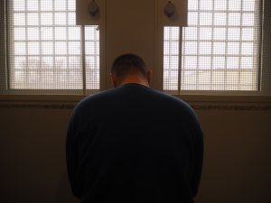 Mężczyzna ze spuszczoną głową stoi tyłem do zdjęcia na tle okien policyjnej celi.