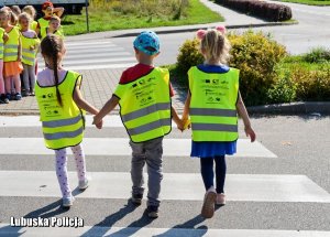 Trójka dzieci trzymając się za ręce przechodzi przez przejście dla pieszych.