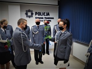 Komendant Powiatowy Policji w Sulęcinie wręcza policjantce różę. W tle widoczni są pozostali policjanci. Wszyscy ubrano są w mundury galowe. Na twarzach mają czarne maseczki ochronne.