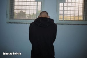 Zatrzymany mężczyzna w czarnej bluzie stoi ze spuszczoną głową tyłem do obiektywu, a przodem do okratowanych okien policyjnej celi.