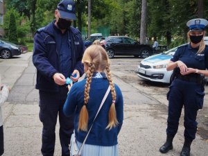 Policjant wraz z policjantką w maseczkach ochronnych wręczają odblask dziewczynce w warkoczykach przed budynkiem szkoły.