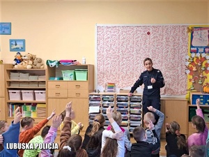 Policjantka prowadzi spotkanie z dziećmi klasy drugiej szkoły podstawowej. Dzieci trzymają ręce do góry aby zadać pytanie. Siedzą tyłem do zdjęcia.