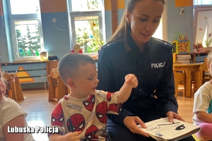 Policjantka siedzi wraz z chłopcem na dywanie w przedszkolnej sali. W rękach trzyma książeczkę, na którą 3-latek wskazuje palcem.