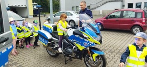Policjant stoi przy motocyklu policyjnym na którym siedzi dziewczynka a obok stoją inne dzieci