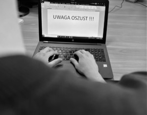 Zakapturzony mężczyzna siedzi przed ekranem laptopa, na którym wyświetlony jest napis: UWAGA OSZUST !!!