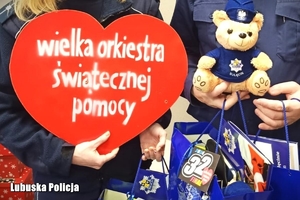 Policjantka trzyma w dłoniach kartonowe serce - symbol Wielkiej Orkiestry Świątecznej Pomocy. Policjant trzyma w dłoniach Misia Sulisia oraz torby z przekazanymi na licytację gadżetami.