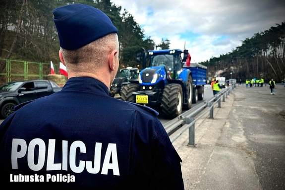 Policjant stoi tyłem do zdjęcia na tle maszyn rolniczych biorących udział w proteście rolników.